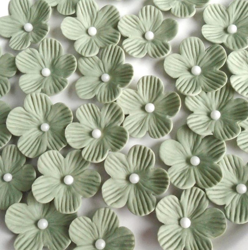 May include: A collection of sage green ceramic flower embellishments with white pearl centers. The flowers have detailed petal textures and are arranged closely together, creating a decorative floral pattern. These are ideal for craft projects.