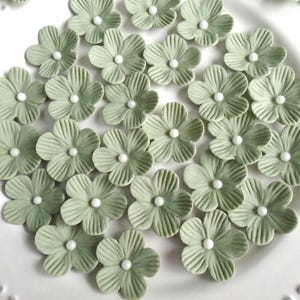 May include: A collection of sage green flower-shaped decorations with white centers, arranged on a white plate. The flowers have detailed petal textures. Ideal for crafting or decorating.