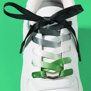 Può includere: Una sneaker bianca con un laccio in nastro di raso nero che sfuma in un gradiente verde. I lacci sono annodati a fiocco nella parte superiore e fissati con un fermalacci metallico color argento a forma di chevron nella parte inferiore.