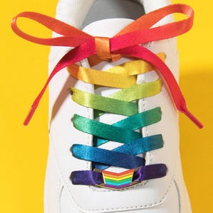 May include: A white trainer featuring flat, satin-like rainbow-coloured shoelaces tied in a bow at the top. A small, rectangular metal charm with a chevron-shaped rainbow pattern is threaded onto the laces near the bottom of the eyelet section.