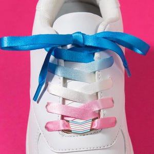 May include: A white sneaker featuring satin shoelaces that transition in color from blue at the top to white in the middle and pink at the bottom. A small metal lace charm with horizontal blue, pink, and white stripes is threaded onto the laces.