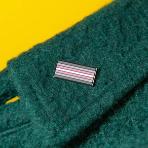 May include: A rectangular enamel pin featuring horizontal stripes in shades of grey, white, and pink, attached to a dark green textured fabric. The pin has a thin black border and is positioned on the folded edge of the thick, fuzzy material.
