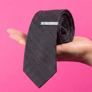 May include: A dark gray necktie with a subtle diagonal weave pattern, featuring a rectangular tie bar with a small horizontal stripe design in light blue, white, and pink colors, held in a rolled position against a bright pink background.