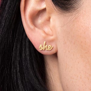 Peut inclure: Une boucle d'oreille à clou dorée en forme du mot she écrit en lettres cursives. La boucle d'oreille est placée sur le lobe de l'oreille, présentant une finition métallique polie et un design en minuscules qui suit la courbe naturelle.
