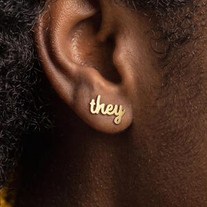 Peut inclure: Une boucle d'oreille à clou dorée en forme du mot they écrit en lettres cursives. La boucle d'oreille est placée sur le lobe de l'oreille, présentant une finition métallique lisse et un design en minuscules qui suit la courbe naturelle.