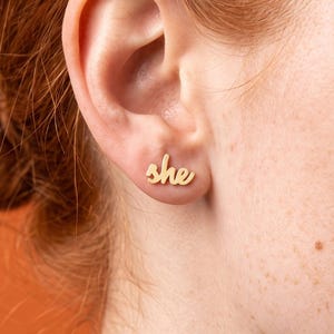 Peut inclure: Une boucle d'oreille à clou dorée en forme du mot she écrit en lettres cursives. Le bijou est placé sur le lobe de l'oreille et présente une finition métallique lisse et polie qui reflète la lumière sur le fond orange uni.