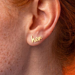 Peut inclure: Une boucle d'oreille à clou dorée en forme du mot her écrit en lettres cursives. La boucle d'oreille est placée sur le lobe de l'oreille et présente une finition métallique lisse et polie qui reflète la lumière, créant un design simple.