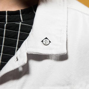 Peut inclure: Une petite épinglette émaillée blanche, de forme circulaire avec une bordure noire et un sommet triangulaire noir. L'épinglette présente une ligne horizontale divisant le centre, avec le texte 01 au-dessus et A1 en dessous, fixée au col d'une veste en jean blanche.