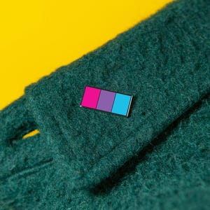 May include: A rectangular enamel pin featuring three vertical stripes in magenta, purple, and cyan, attached to a dark green textured fabric. The pin has a thin metallic border and is positioned on the folded edge of the thick, fuzzy material.