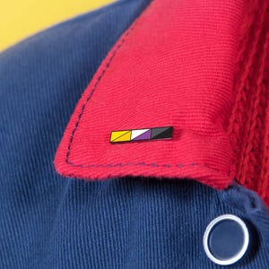 Könnte beinhalten: Eine rechteckige Emaille-Anstecknadel mit einem horizontalen Streifenmuster in Gelb, Weiß, Violett und Schwarz. Die Anstecknadel ist am roten Kordkragen einer marineblauen Jacke befestigt, die über einem leuchtend roten Zopfmuster-Pullover getragen wird.