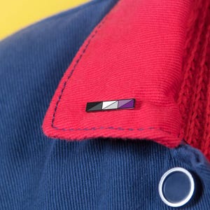 May include: A rectangular enamel pin featuring a horizontal design with black, white, gray, and purple diagonal stripes. The pin is attached to the red collar of a blue jacket, which is worn over a red cable knit sweater with a round button.
