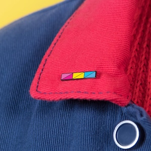 May include: A small rectangular enamel pin featuring three diagonal colour blocks in magenta, yellow, and cyan, attached to the red corduroy collar of a navy blue jacket. The pin has a thin black metal border and is positioned on the right side.