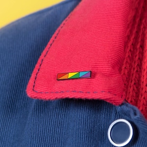 May include: A small rectangular enamel pin featuring a horizontal row of four triangles in red, yellow, green, and blue, attached to the red corduroy collar of a navy blue jacket worn over a red knit jumper.