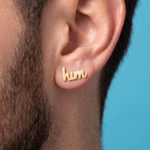 Peut inclure: Une boucle d'oreille à clou dorée en forme du mot hum écrit en lettres cursives minuscules. La boucle d'oreille est placée sur le lobe de l'oreille, présentant une finition métallique lisse et un design épuré qui suit le contour de l'oreille.