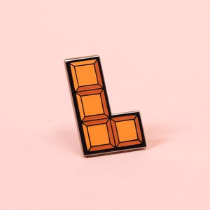 May include: An L-shaped enamel pin featuring a bright orange color with a segmented block design. The pin has a thin black metal outline surrounding the orange sections, creating a distinct geometric shape reminiscent of a classic video game piece.