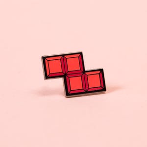 May include: A red enamel pin shaped like a Z-block tetromino, consisting of four connected squares with a dark metal outline. The design features a flat, smooth surface with a vibrant red color and a distinct border separating each individual square.