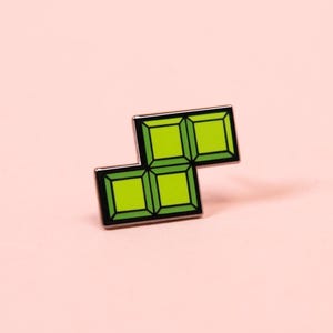 May include: A lime green enamel pin shaped like a four-block geometric puzzle piece. The pin features a black metal outline and internal grid lines that divide the shape into four distinct squares, each with a slightly lighter green center.