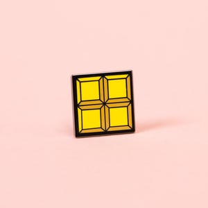 May include: A square enamel pin featuring a bright yellow grid design divided into four smaller squares by thin black lines. The pin has a distinct black border framing the yellow segments, creating a clean and geometric appearance.
