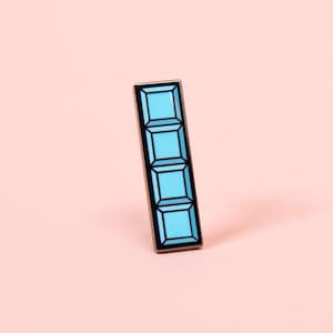 Puede incluir: Un pin de esmalte rectangular con un diseño vertical de cuatro segmentos cuadrados de color azul claro apilados, delineados con metal negro. El pin presenta una estética geométrica limpia con un acabado liso y un borde de metal oscuro.