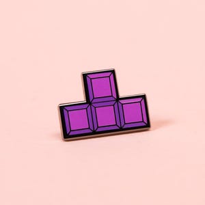 May include: A purple enamel pin shaped like a T-block tetromino. The pin features four square segments arranged in a T-shape, each with a darker purple border and a lighter purple center, outlined in a thin metallic frame against a solid background.