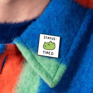 Può includere: Una spilla smaltata rettangolare con sfondo bianco, un'illustrazione di una rana verde in stile cartoon e il testo nero "STATUS: TIRED" stampato sopra e sotto la rana. La spilla è fissata su una giacca a righe blu, verdi e arancioni.