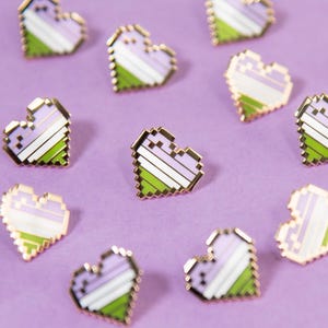 Könnte beinhalten: Eine Sammlung kleiner herzförmiger Emaille-Anstecknadeln mit pixeligem Design. Jede Anstecknadel zeigt horizontale Streifen in hellen Violett-, Weiß- und Grüntönen, wobei ein metallisch goldfarbener Rand die Herzform deutlich hervorhebt.