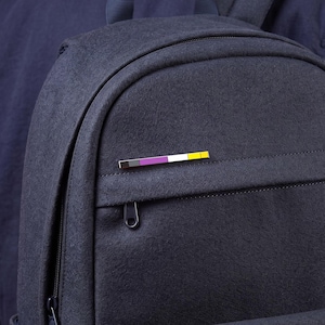 May include: A dark navy blue felt backpack featuring a horizontal rectangular enamel pin attached to the front pocket. The pin displays a color sequence of black, gray, white, purple, and yellow stripes arranged in a thin, elongated bar shape.
