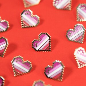 Puede incluir: Una colección de pequeños pines de esmalte con forma de corazón pixelado dispuestos de forma dispersa. Cada pin presenta un diseño a rayas con bandas horizontales de color blanco, rosa claro, rosa medio y rosa oscuro, con un borde dorado.