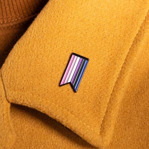 May include: A rectangular enamel pin with a notched bottom edge, featuring vertical stripes in shades of pink, white, purple, black, and blue, attached to the collar of a mustard yellow wool coat worn over a brown ribbed turtleneck sweater.