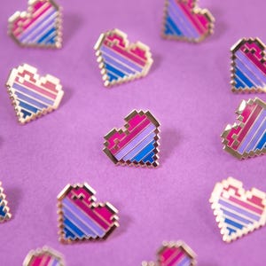 May include: A collection of small, pixelated heart-shaped enamel pins. Each pin features a striped pattern of pink, purple, and blue colours with a gold-toned metal outline. The hearts are arranged in a scattered pattern across the frame.