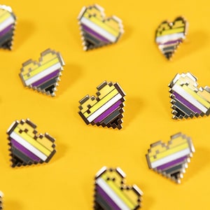 May include: A collection of small enamel pins shaped like pixelated hearts. Each heart features horizontal stripes in yellow, white, purple, and black. The pins are arranged in a scattered pattern, showcasing the distinct blocky, digital-style edges.