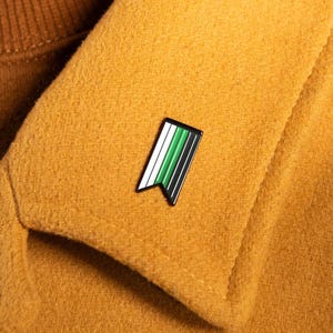 Puede incluir: Un pequeño pin de esmalte rectangular con borde inferior en forma de cola de golondrina, con rayas verticales en blanco, verde claro, verde oscuro y negro. El pin está sujeto a la solapa de un abrigo amarillo mostaza sobre un cuello alto marrón.