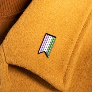 May include: A small, rectangular enamel pin shaped like a pennant flag with a black metal border. The pin features vertical stripes in light purple, white, and green, attached to the collar of a mustard yellow coat worn over a brown turtleneck.