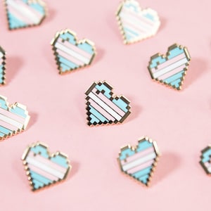 May include: A collection of small enamel pins shaped like pixelated hearts. Each heart features horizontal stripes in light blue, white, and pink, outlined in a gold-toned metal border. The pins are arranged in a scattered pattern across the frame.