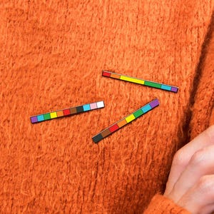 Puede incluir: Tres pines rectangulares de esmalte con rayas horizontales de varios colores. Un pin muestra una secuencia de arcoíris estándar, mientras que los otros dos incorporan segmentos adicionales en marrón, negro, blanco, rosa y azul claro.