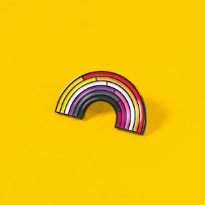 Könnte beinhalten: Eine Metall-Emaille-Anstecknadel in Form eines Regenbogenbogens. Das Design zeigt horizontale Streifen in einem Farbverlauf aus Rot, Orange, Gelb, Weiß, Lila und Dunkelgrau, die alle von dünnen schwarzen Metallrändern auf gelbem Grund umrandet sind.