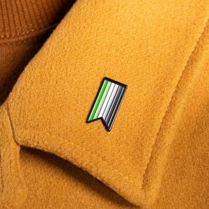 May include: A small, rectangular enamel pin with a swallowtail bottom edge, featuring vertical stripes in shades of light green, white, gray, and black, attached to the lapel of a mustard yellow wool coat worn over a brown turtleneck sweater.