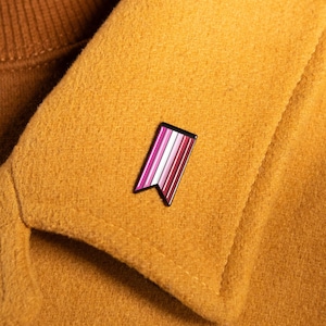 Puede incluir: Un pequeño pin de esmalte rectangular con forma de estandarte vertical y borde inferior dentado. El pin presenta una serie de rayas verticales en tonos rosa, blanco y rojo oscuro, todo ello encerrado en un fino borde de metal oscuro.