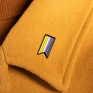 May include: A metal enamel pin shaped like a vertical banner with a swallowtail bottom edge. The pin features four vertical stripes in yellow, white, purple, and black, outlined in a thin black metal border, attached to a mustard yellow coat collar.