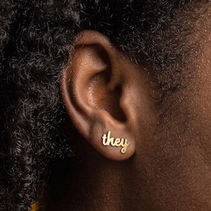 May include: A gold-toned stud earring shaped like the word they in a cursive script font. The earring is positioned in the earlobe, featuring a smooth, polished metallic finish that reflects light, creating a clear and legible text design.