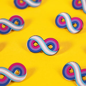 May include: Multiple enamel pins shaped like the infinity symbol. Each pin features a striped pattern of pink, purple, and blue colors with a white center stripe. The pins are arranged in a repeating pattern across the frame, showcasing the design.