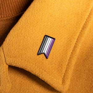 May include: A small, rectangular metal enamel pin shaped like a vertical banner with a notched bottom edge. The pin features four vertical stripes in black, gray, white, and purple, enclosed within a thin black metal border, attached to a mustard coat.