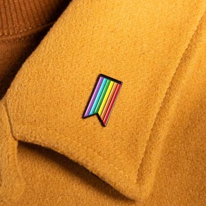 Könnte beinhalten: Eine rechteckige Emaille-Anstecknadel mit spitz zulaufender Unterkante, die vertikale Streifen in Rot, Orange, Gelb, Grün, Blau und Violett aufweist und am Revers eines senfgelben Mantels über einem braunen Rollkragenpullover befestigt ist.