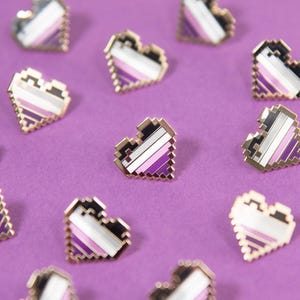 May include: A collection of small, pixelated heart-shaped enamel pins. Each pin features a diagonal striped pattern in black, grey, white, and purple colors, with a metallic gold-toned border outlining the jagged, blocky edges of the heart shape.