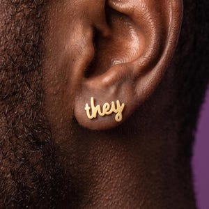 Peut inclure: Une boucle d'oreille à clou en métal doré en forme du mot they écrit en lettres cursives. La boucle d'oreille est placée sur le lobe de l'oreille, présentant une surface lisse et polie qui reflète la lumière avec une écriture claire.