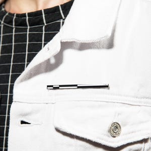May include: A white denim jacket featuring a black and white grid pattern shirt underneath. A rectangular enamel pin with a black and white scale pattern is attached to the chest pocket area of the jacket, which has silver metal buttons.