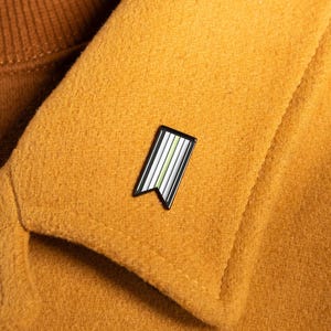 May include: A rectangular enamel pin with a swallowtail bottom edge, featuring vertical stripes in shades of white, light green, and dark green, outlined in black. The pin is attached to the lapel of a mustard yellow coat worn over a brown jumper.