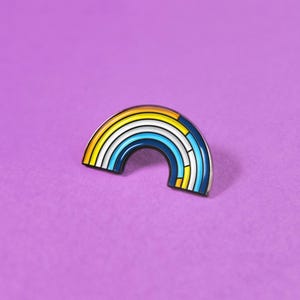 May include: A metal enamel pin shaped like a semi-circular rainbow. The design features concentric bands of yellow, orange, light blue, white, and dark blue. The right side of the rainbow includes vertical segments that break up the color bands.