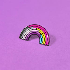 May include: A metal enamel pin shaped like a rainbow arch. The design features horizontal segments of color including dark gray, light gray, white, pink, purple, blue, and yellow, all outlined in black metal. The pin has a smooth, glossy finish.