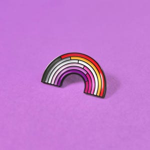 May include: A metal enamel pin shaped like a rainbow arch. The design features seven concentric bands of color, transitioning from dark red at the outer edge through orange, yellow, white, light pink, and dark pink to a deep purple at the center.